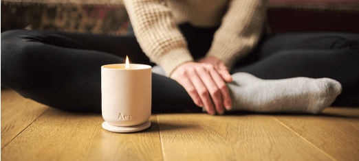 figure meditating in comfy clothes on a wooden floor next to a lit candle