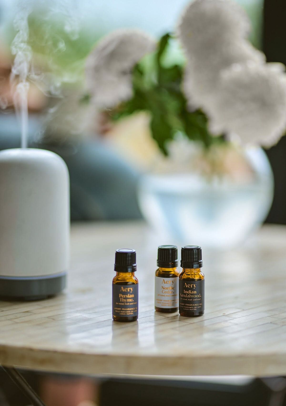 White Wonderland set of three fragrance oils by Aery displayed next to electric diffuser and vase of flowers on round table