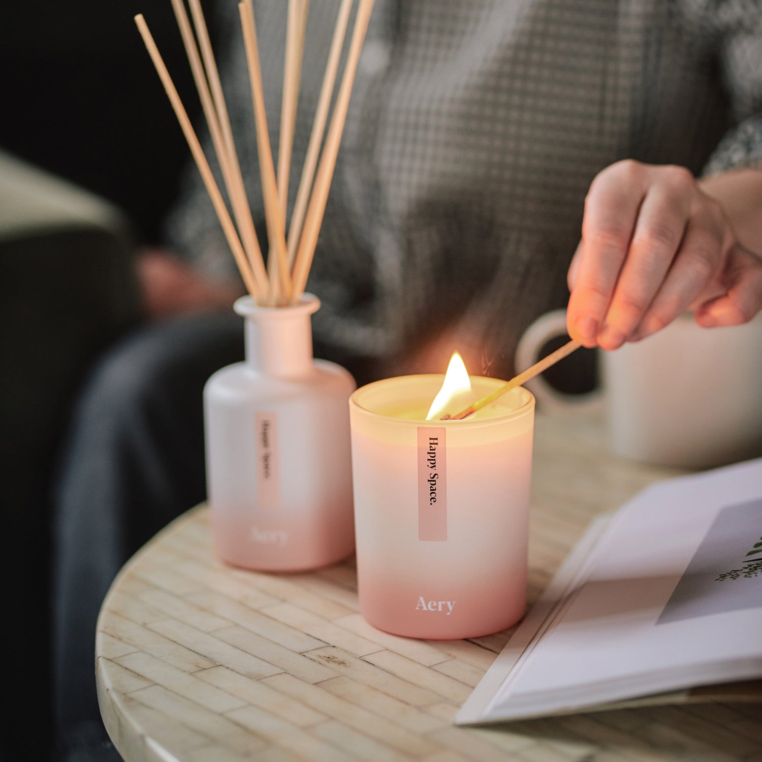 persons hand lighting a happy space candle displayed next to matching diffuser on coffee table