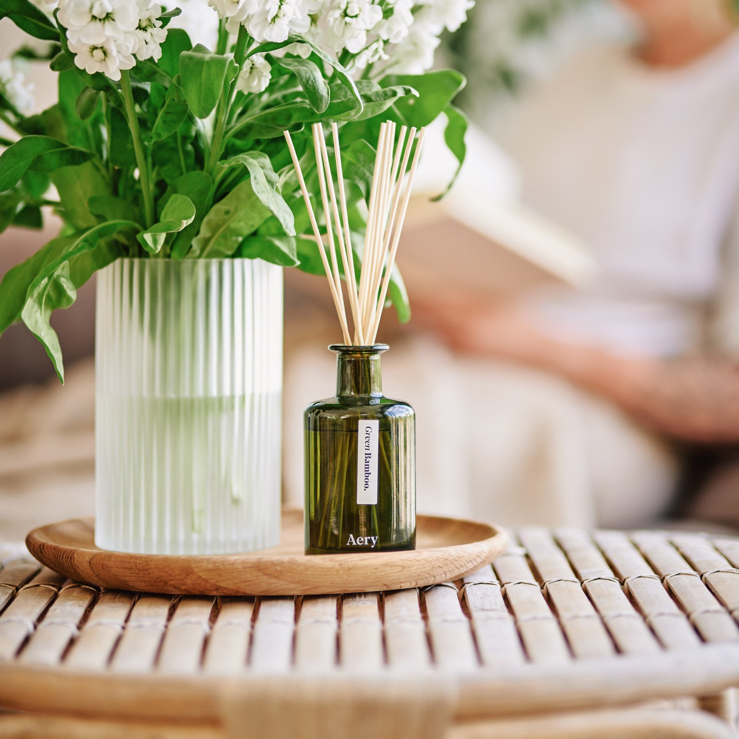 Green bamboo botanical diffuser displayed on a coffee table next to a vase filled with fresh white stocks