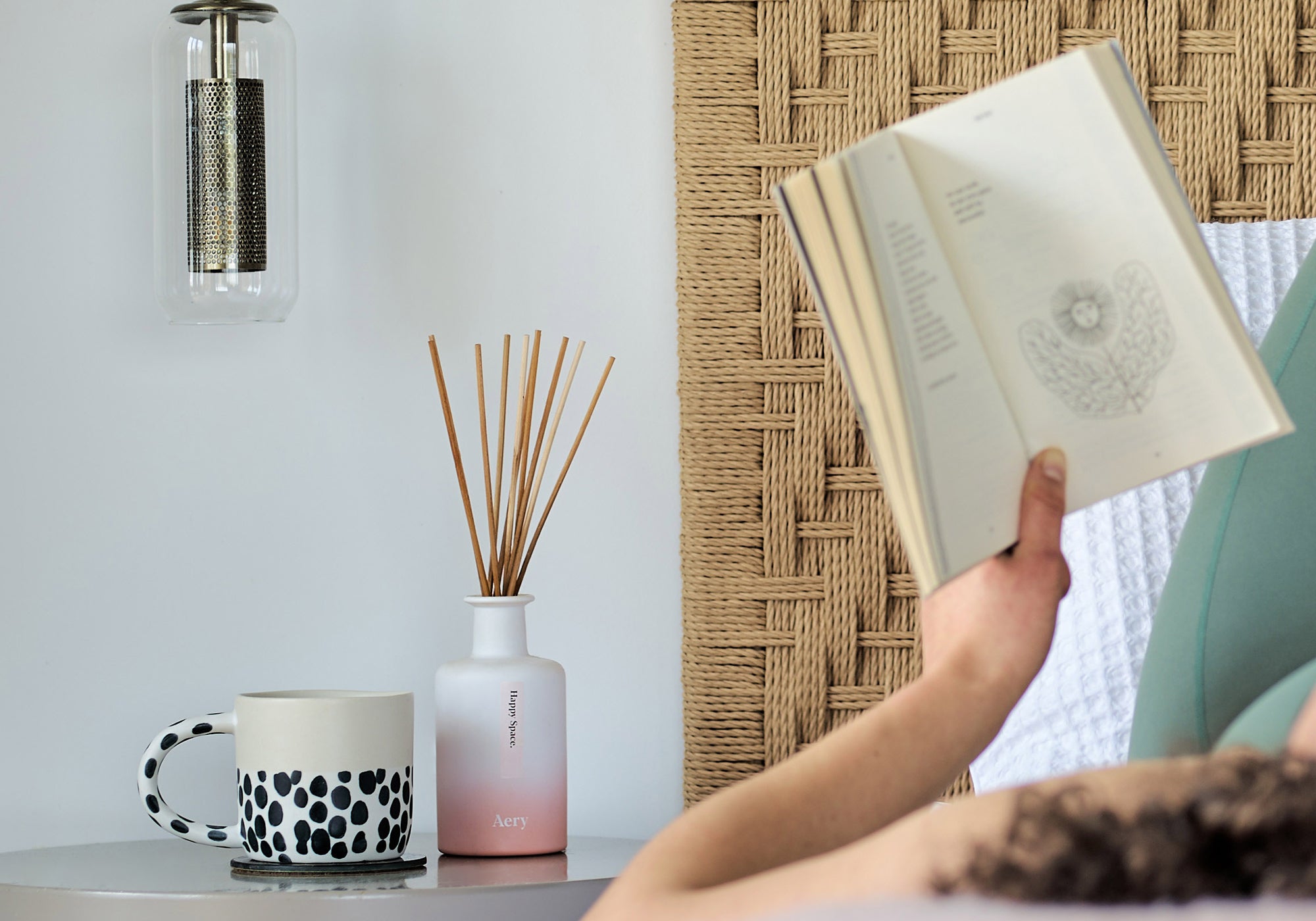 pink happy space reed diffuser on bedside table next to coffee cup with person reading in the foreground