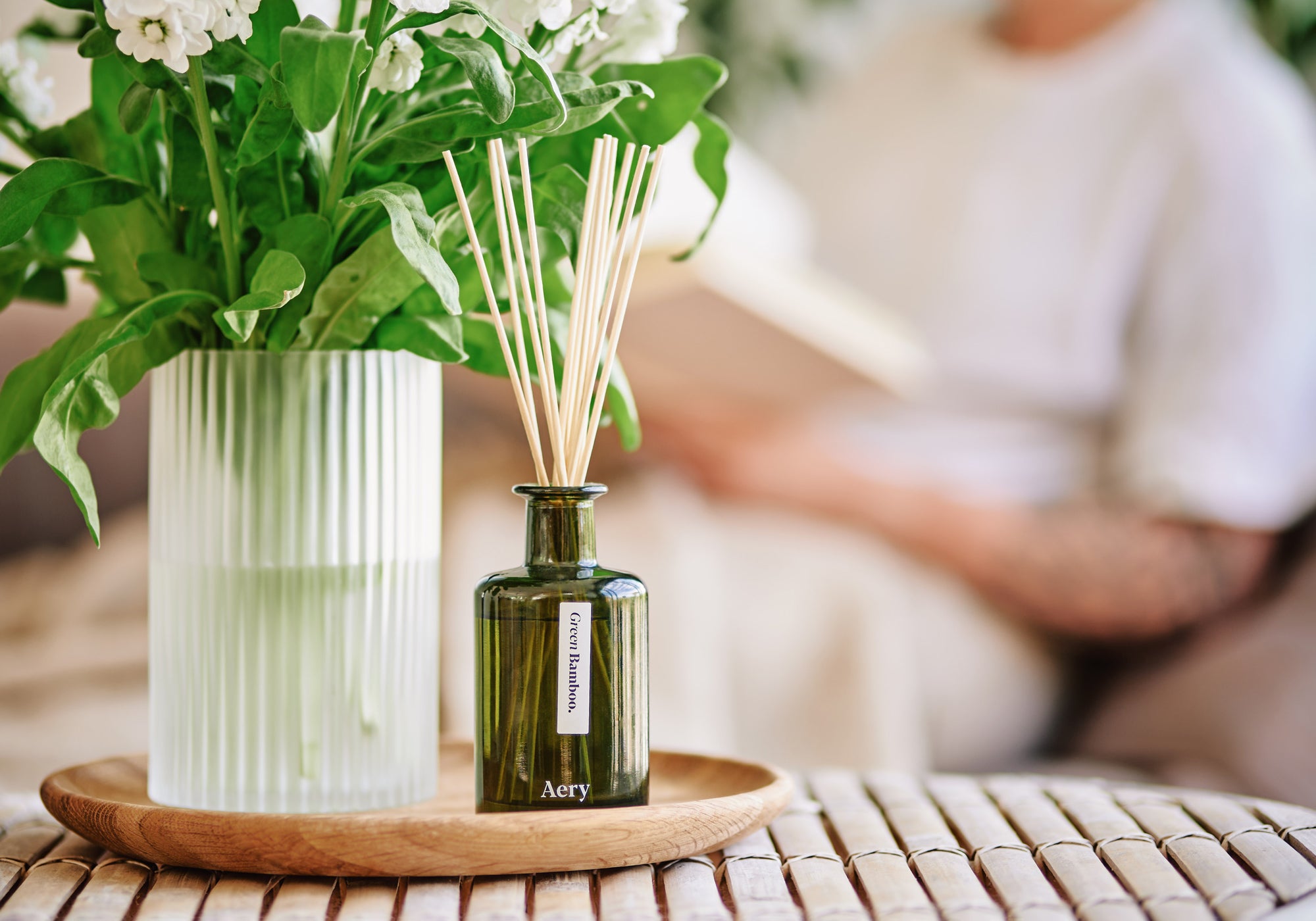 green glass diffuser displayed on a coffee table next to a bunch of white flowers with person reading in the background