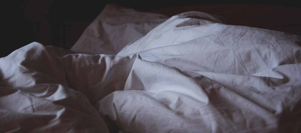 Photo of a crumbled white bed spread and pillow in a dimly lit rook