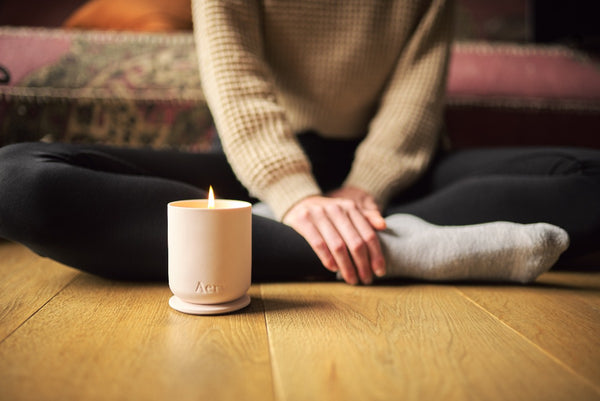 figure meditating in comfy clothes on a wooden floor next to a lit candle