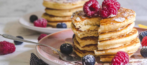 stack of pancakes surrounded by maple syrup, blueberries and raspberries. 