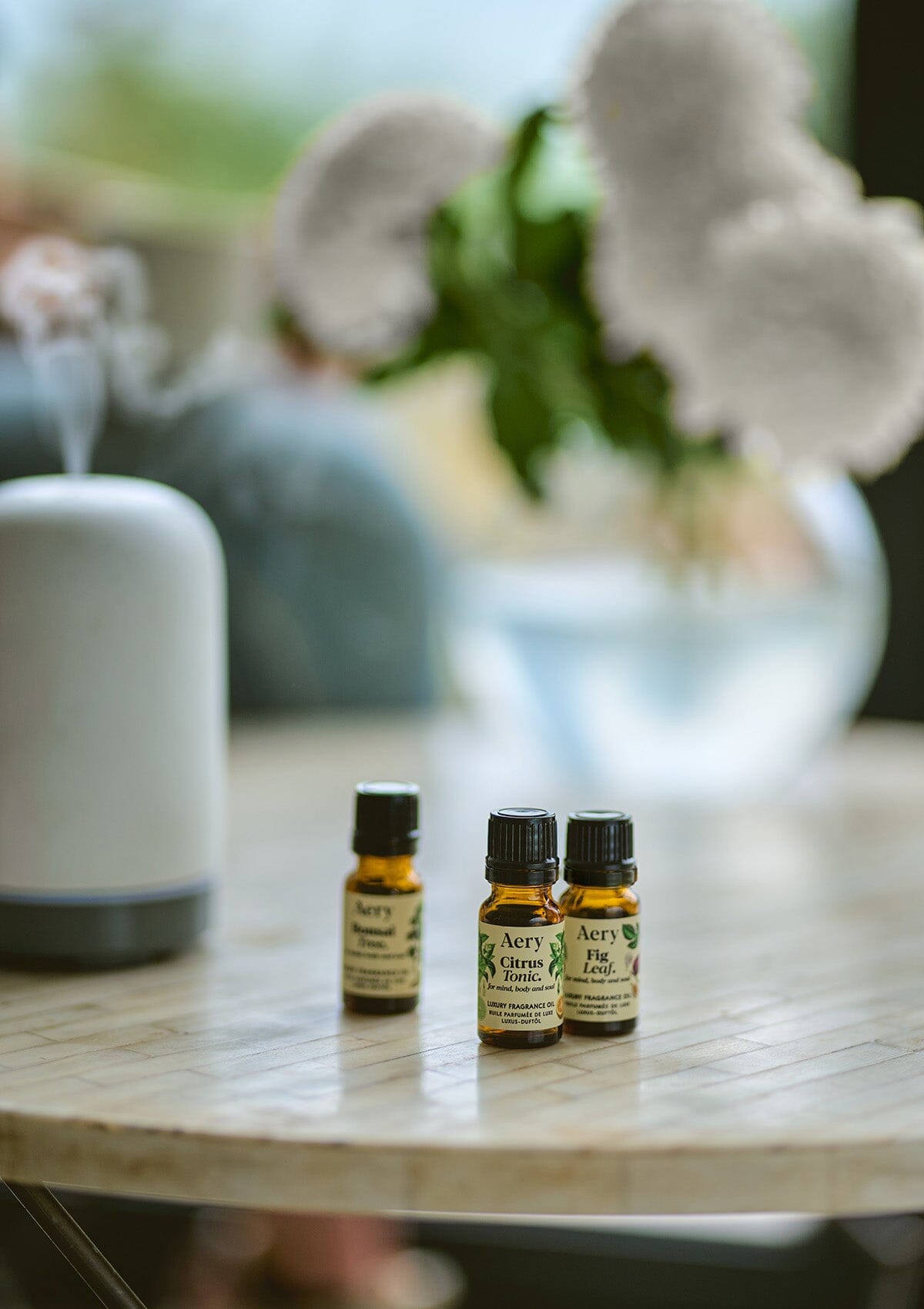 Cream Botanical set of three fragrance oils by Aery displayed next to electric diffuser and vase of flowers on round table 