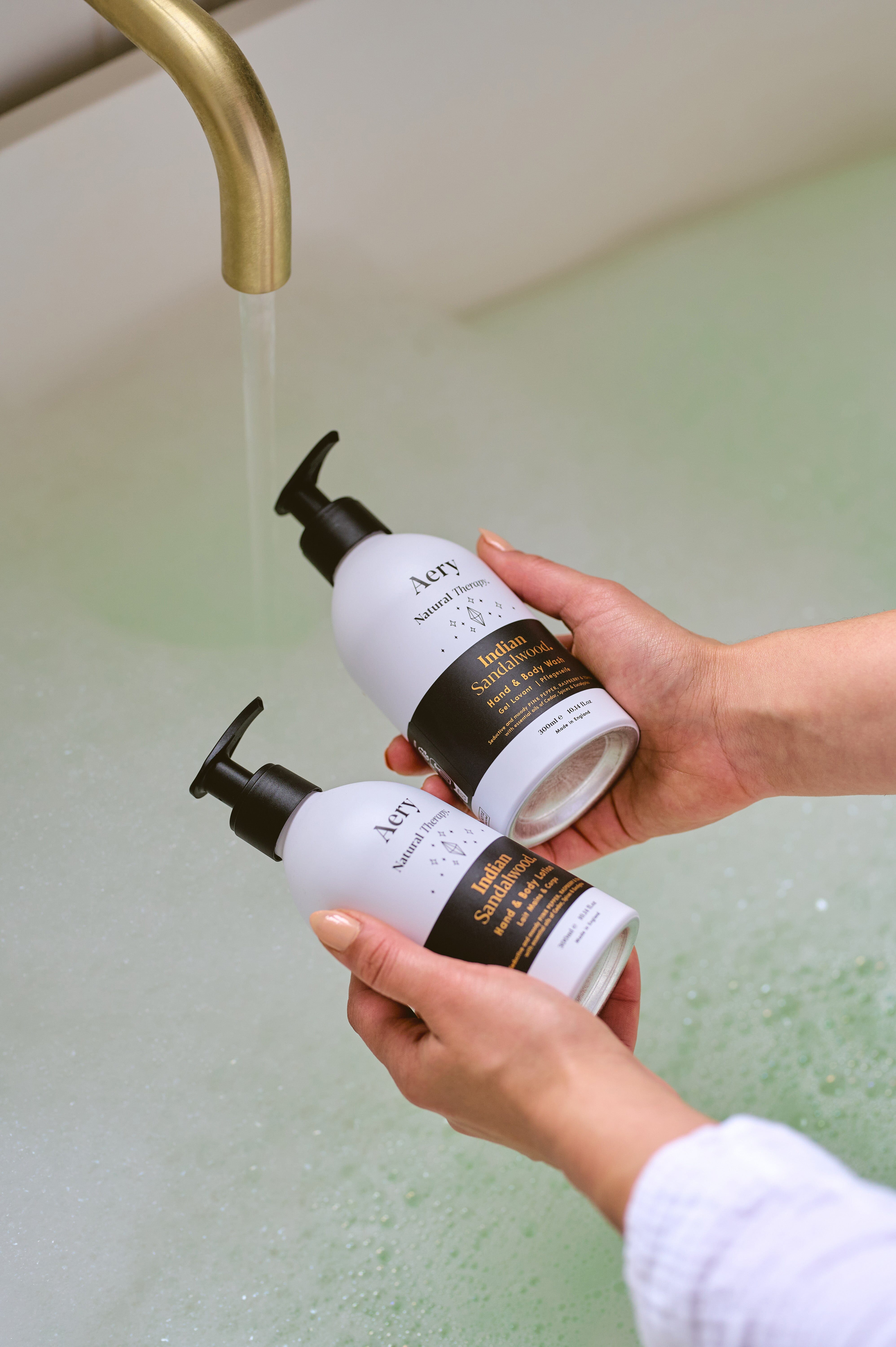 Black Indian Sandalwood hand and body wash and lotion duo by Aery displayed in hands over bubble bath 