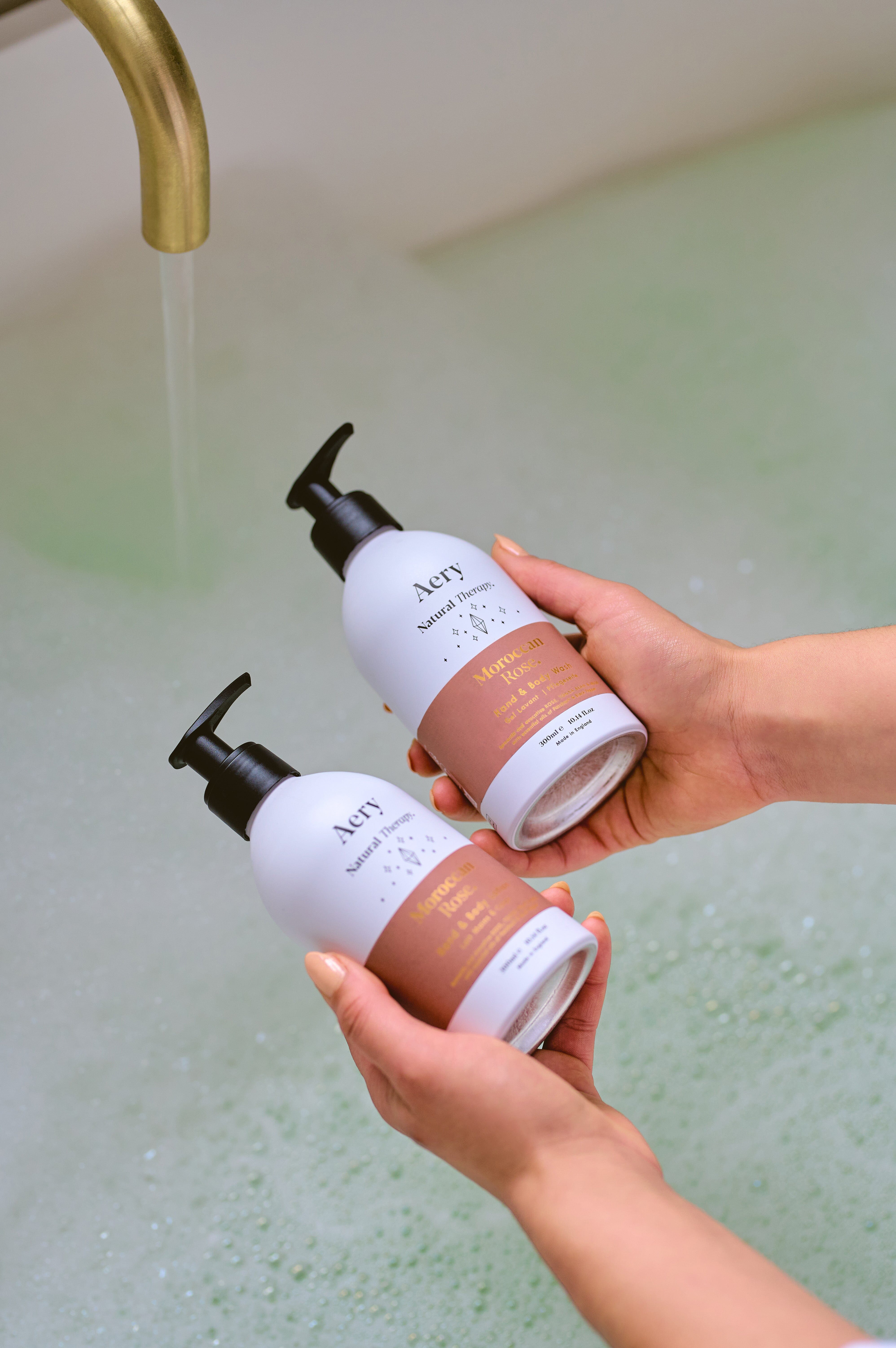 Aubergine Moroccan Rose hand and body wash and lotion duo by Aery displayed in hands over bubble bath 