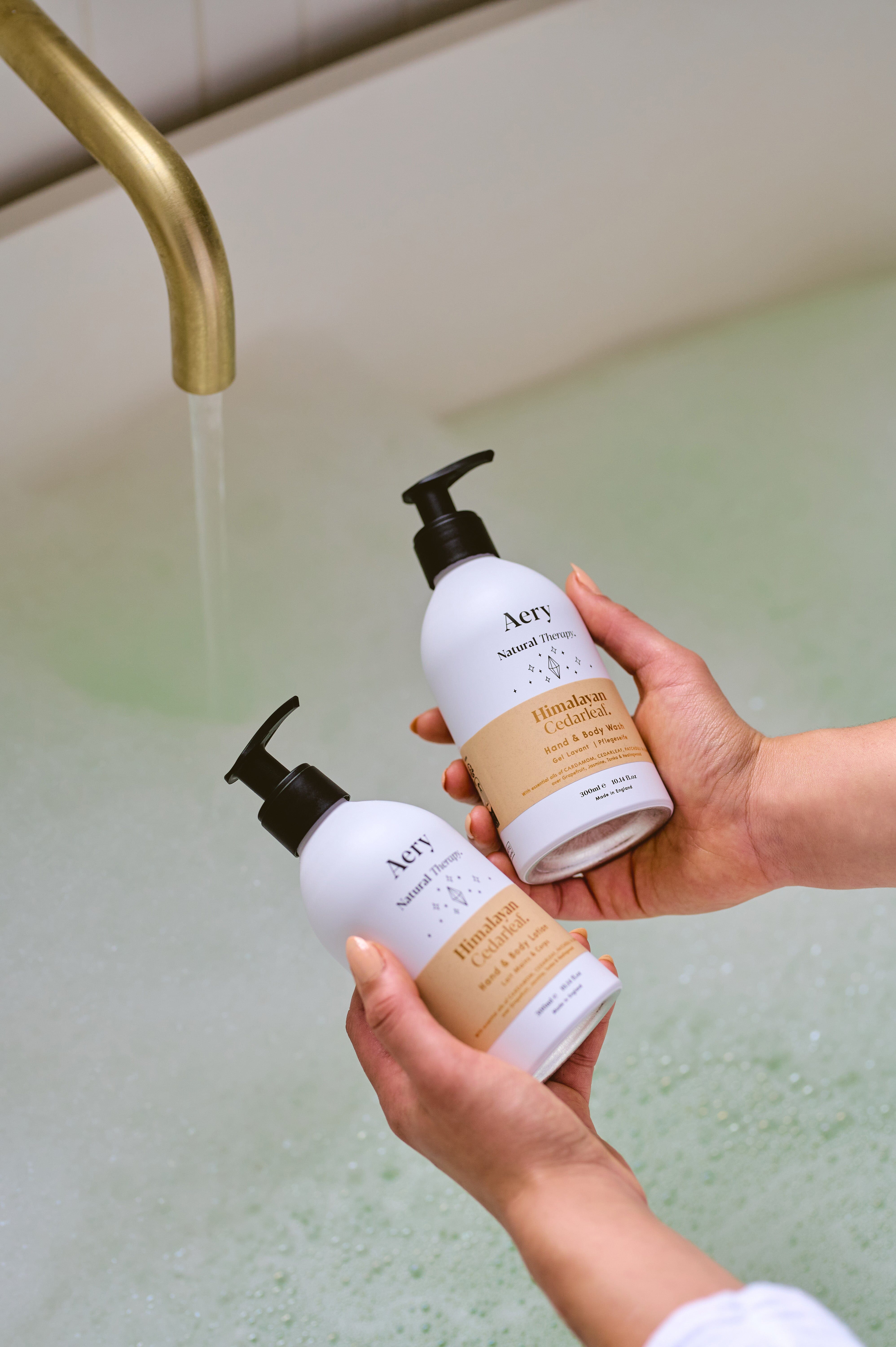 Cream Himalayan Cedarleaf hand and body and lotion and wash duo by Aery held in hands over bubble bath 