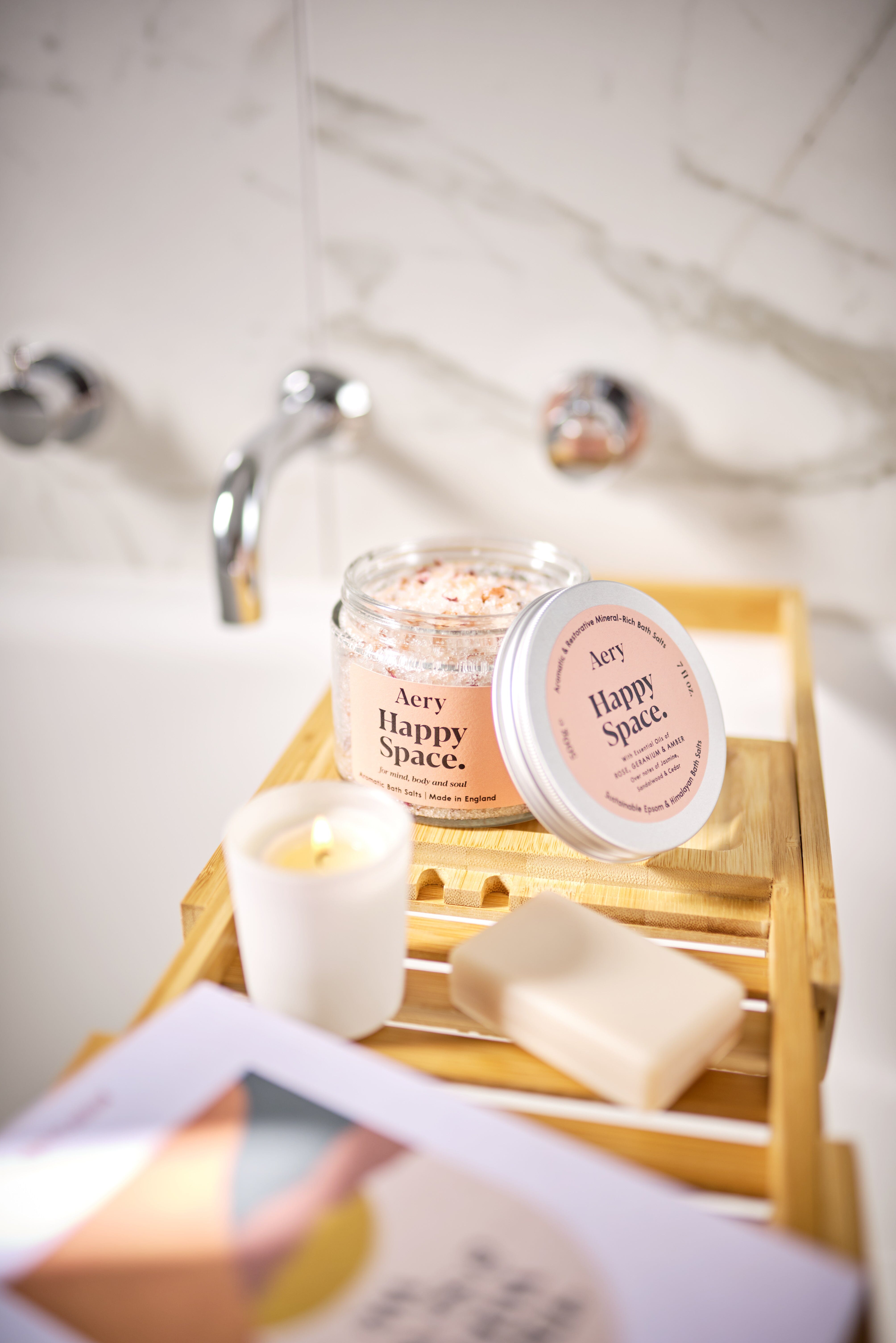 Pink Happy Space baths salts jar by Aery displayed next to soap bar and mini candle on wooden bath tray in bathroom 