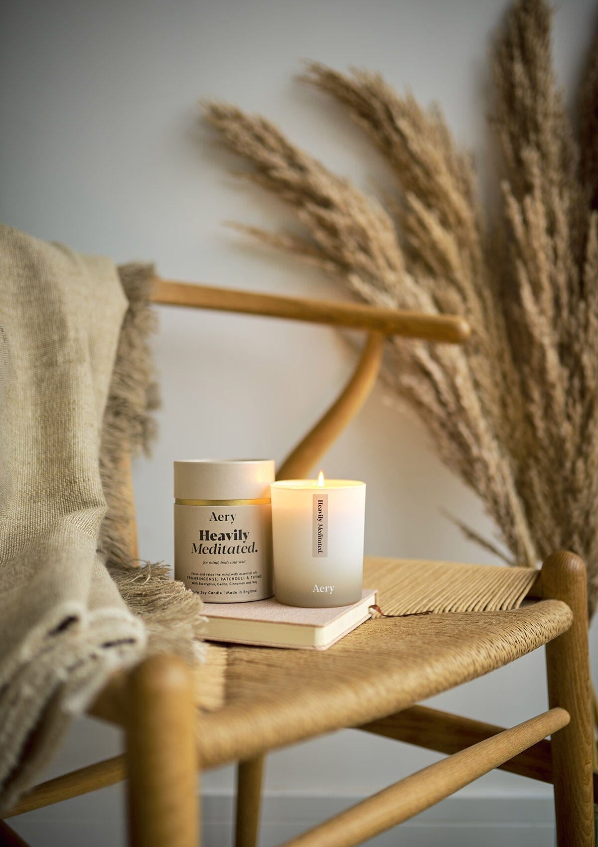 Beige Heavily Meditated candle displayed next to product packaging by Aery placed on book and wicker chair 