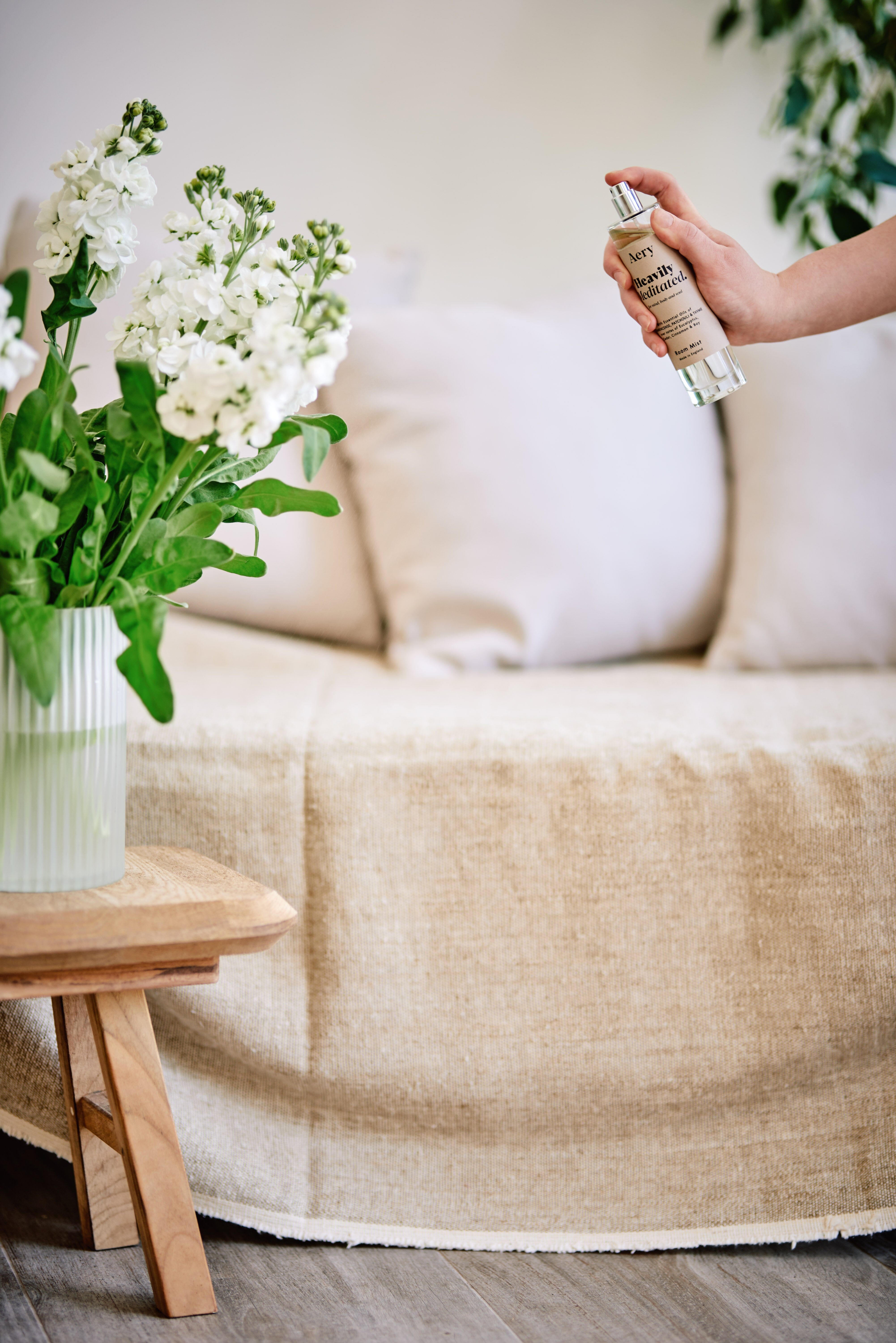 Beige Heavily Meditated room mist by Aery displayed in hand next to vase of white flowers 