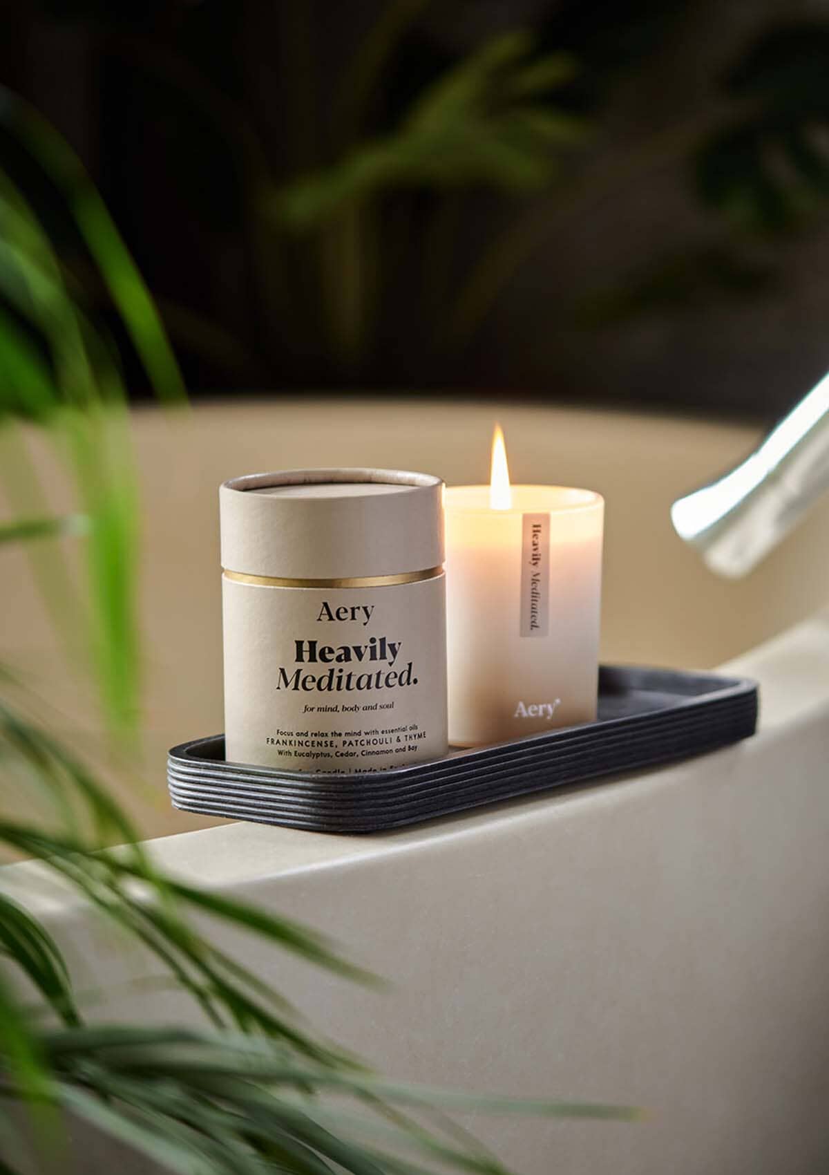 Beige Heavily Meditated Candle displayed next to product packaging by Aery placed on black tray in bathroom 