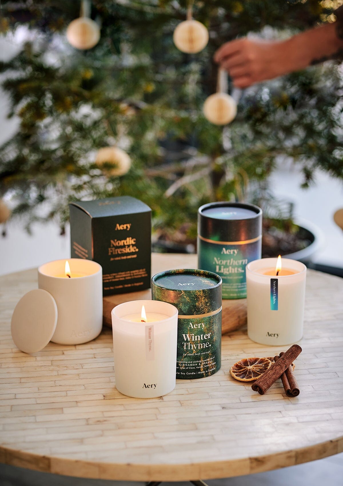 Winter Thyme Nordic Fireside and Northern Lights candles by Aery displayed next to product packaging placed on cream circle table in front of Christmas tree 