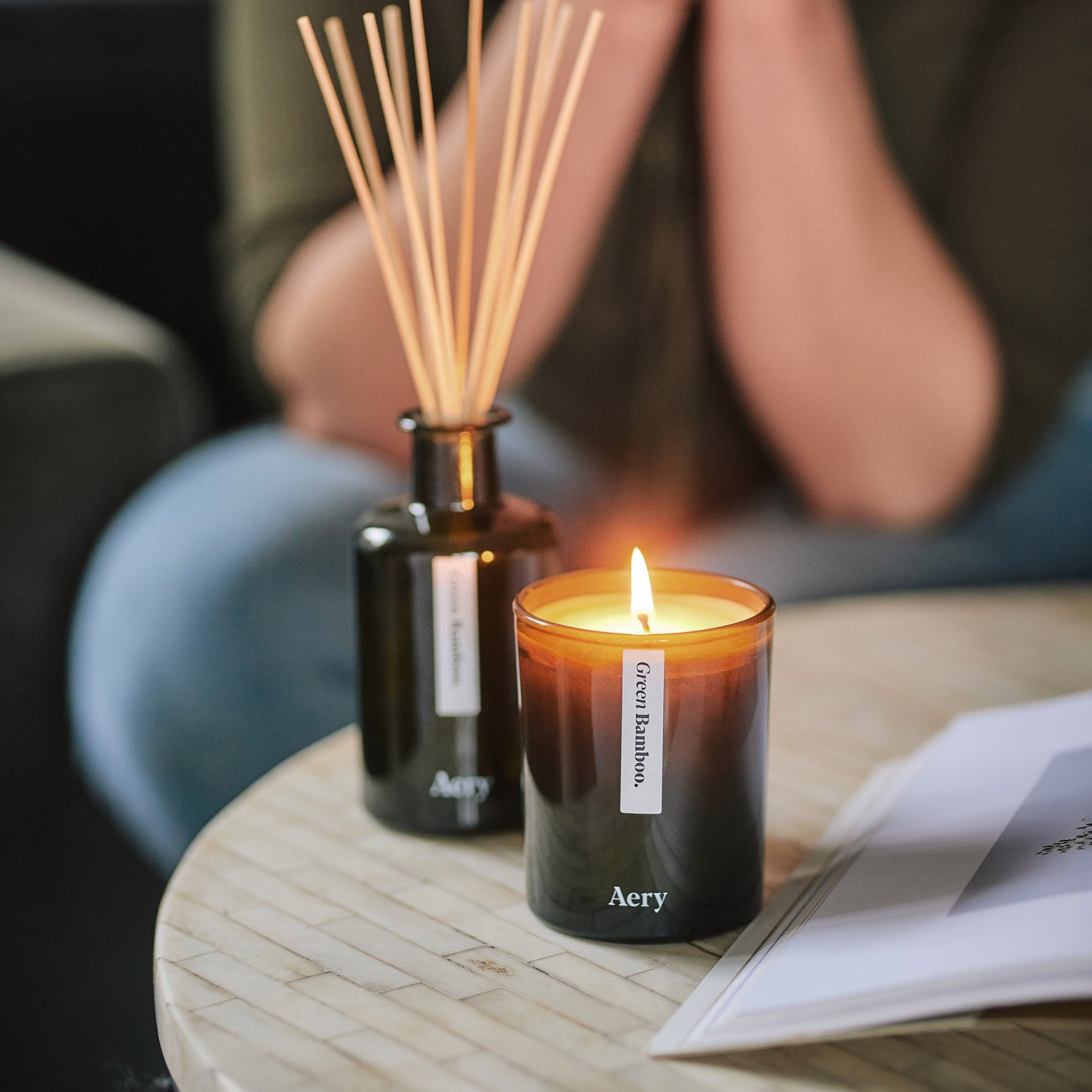 Person sat at coffee table with magazine with lit green bamboo candle and matching diffuser