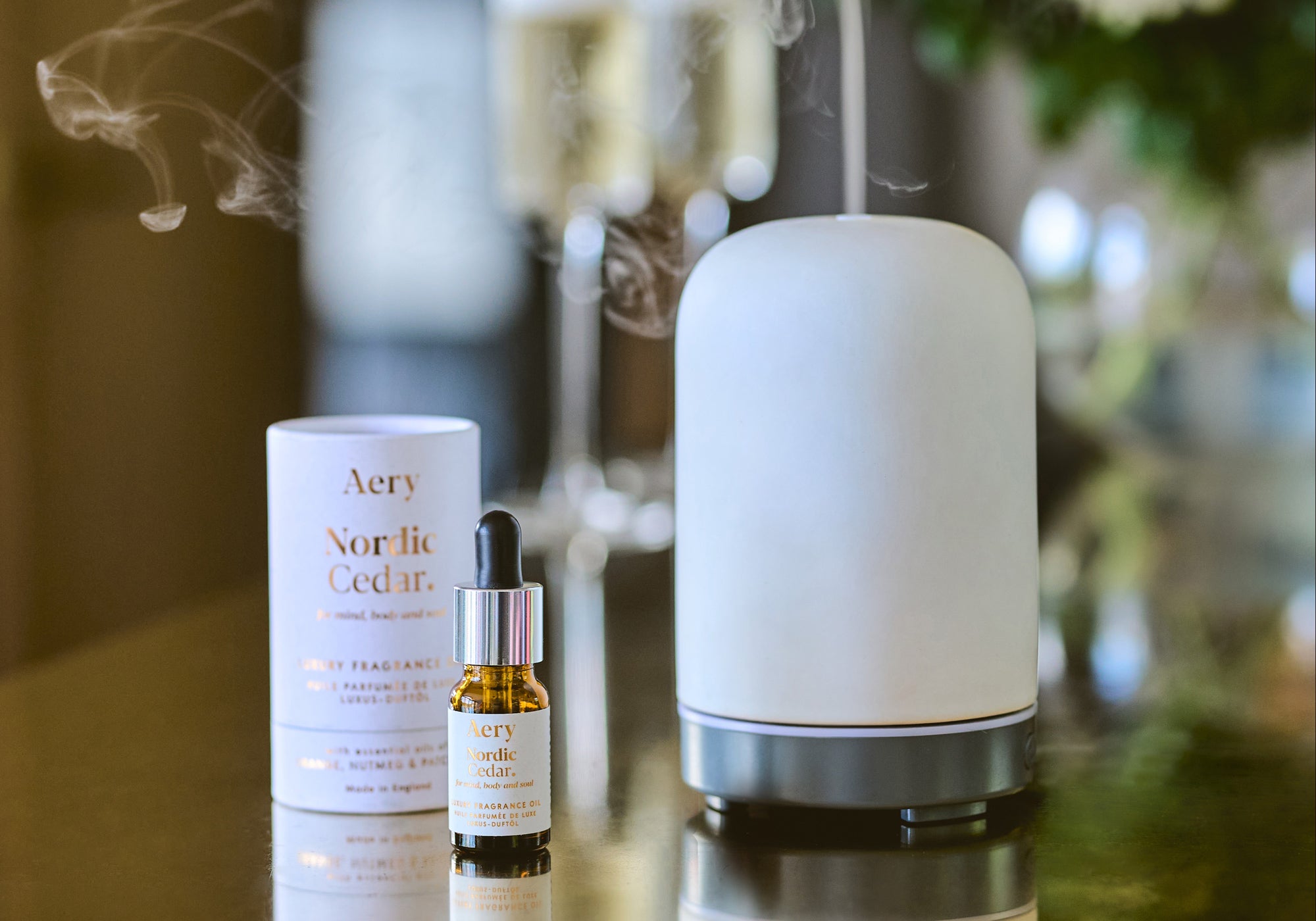 nordic cedar air diffuser oil displayed next to air diffuser and product packaging with flowers and prosecco glasses in the background