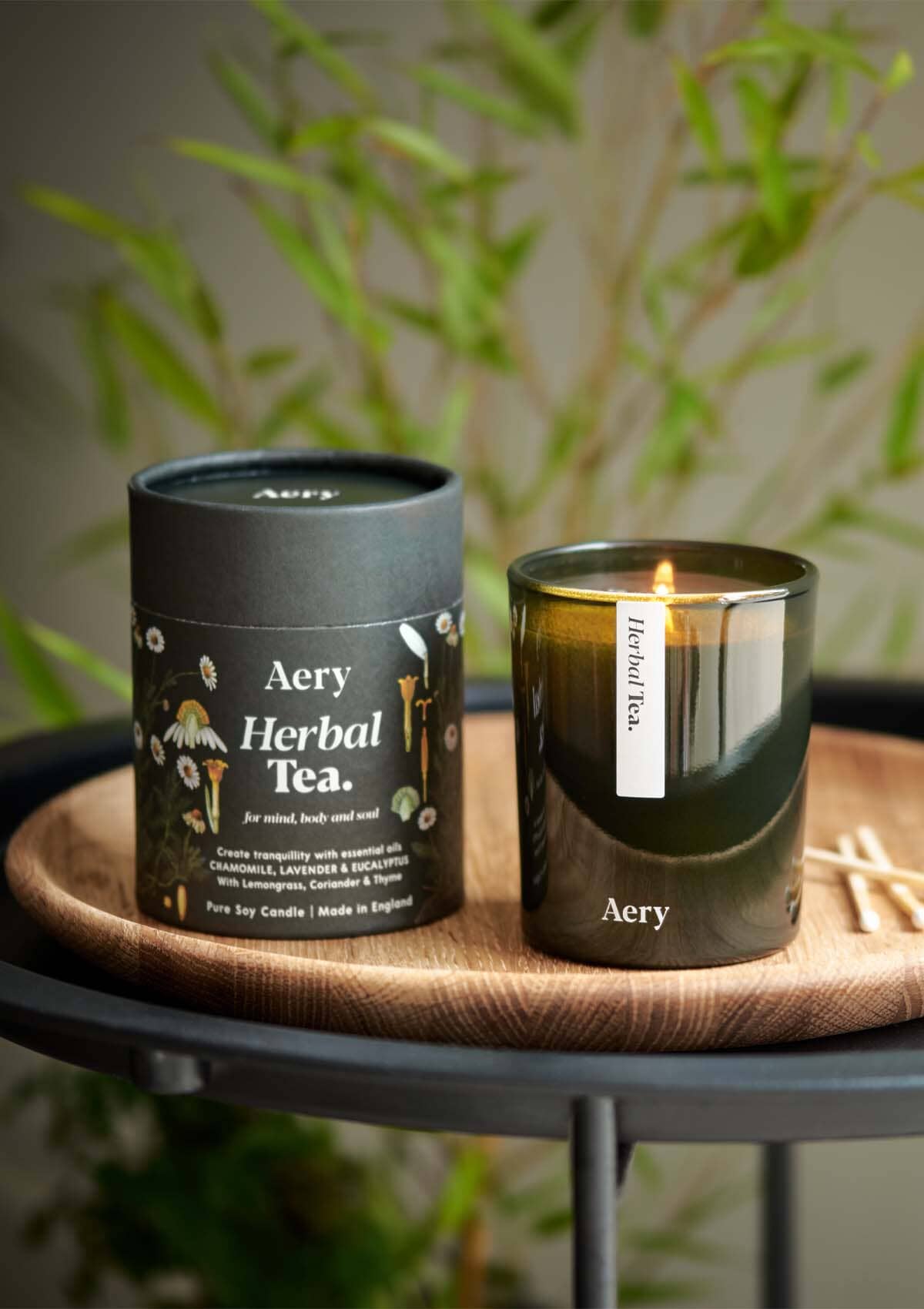 Green Herbal Tea candle displayed next to product packaging by Aery placed on wooden tray with green plant behind 