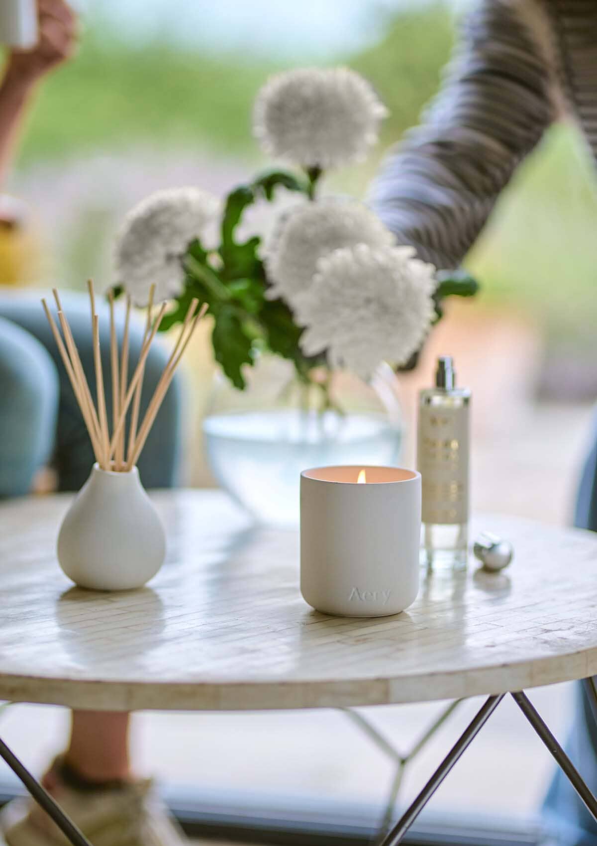 White Nordic Cedar candle by Aery displayed next to nordic cedar diffuser and room mist and vase of white flowers placed on cream circle table