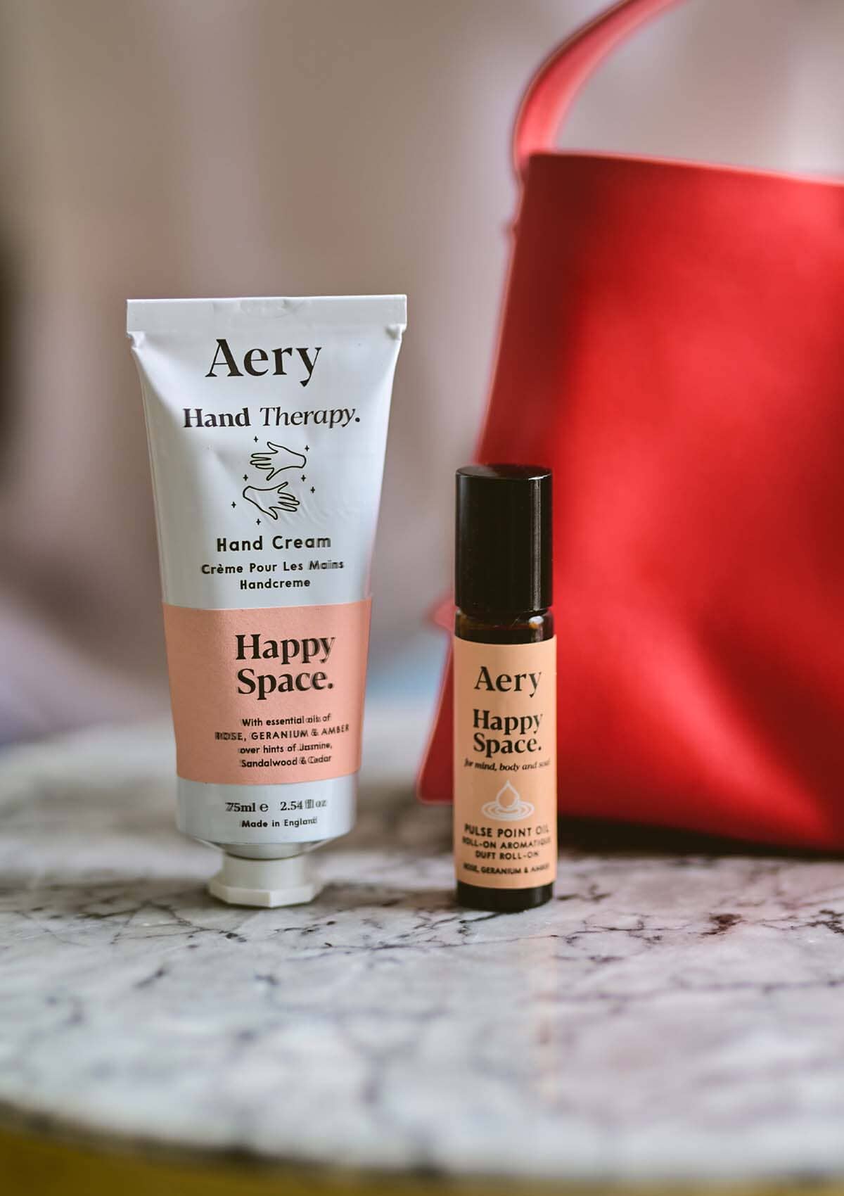 Pink Happy Space pulse point oil and Happy Space hand cream by Aery in front of red hand bag 