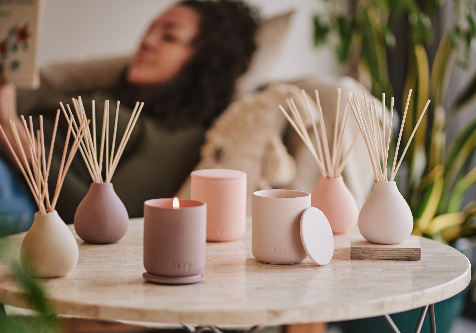 Person laying in background reading a book whilst pink clay candles and diffusers by aery living are displayed on a coffee table