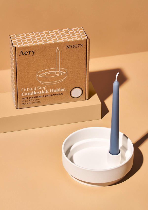 aery white ceramic candle holder with grey tapered candle next to kraft box packaging against an orange background