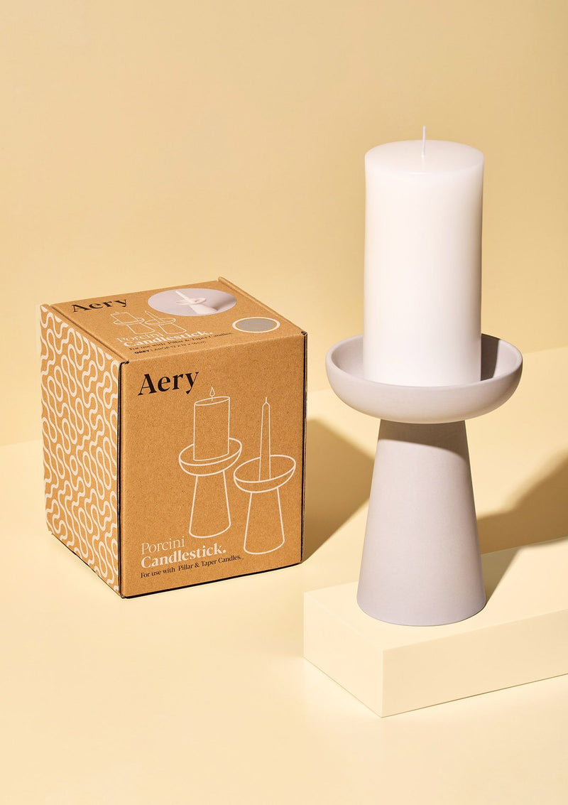 aery living tall grey ceramic candle holder displayed with a large white pillar candle and next to product packaging against a yellow background