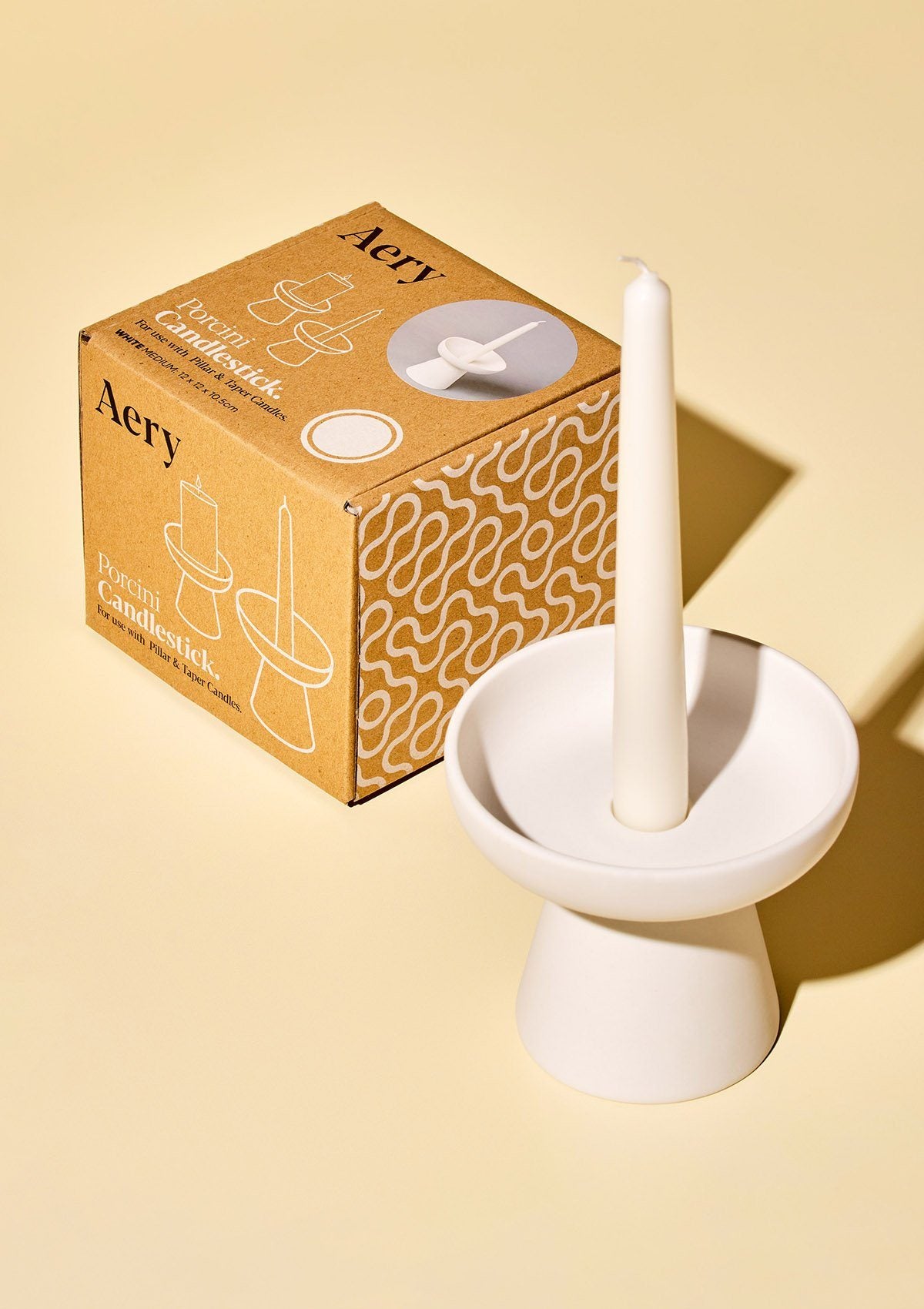 aery living white ceramic candle holder displayed with tapered white candle next to product packaging against a yellow background